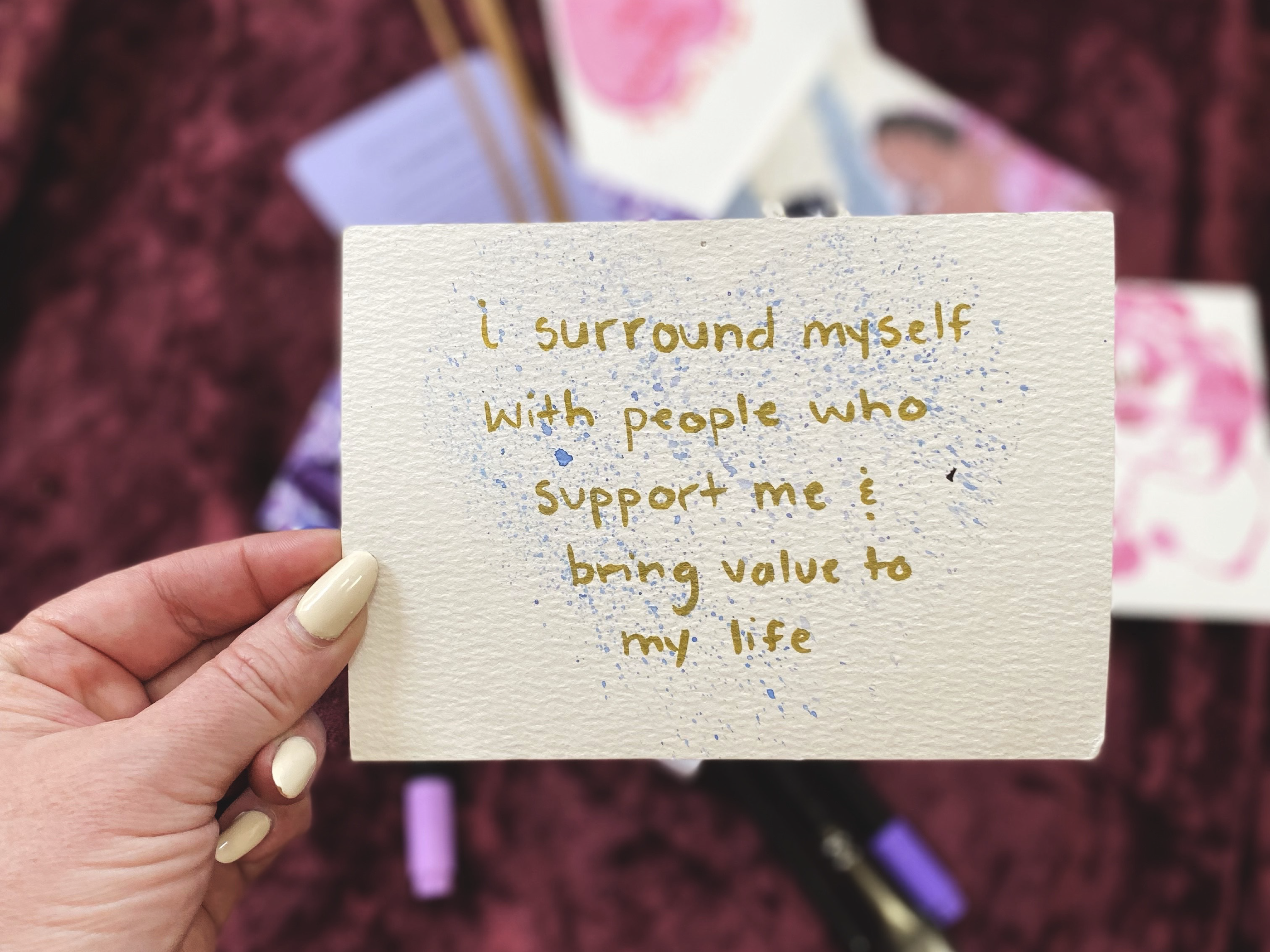 A women's hand with butter yellow painted nails, holds up a card that reads "I surround myself with people who support me & bring value to my life". In the background, various art supplies cover the table.