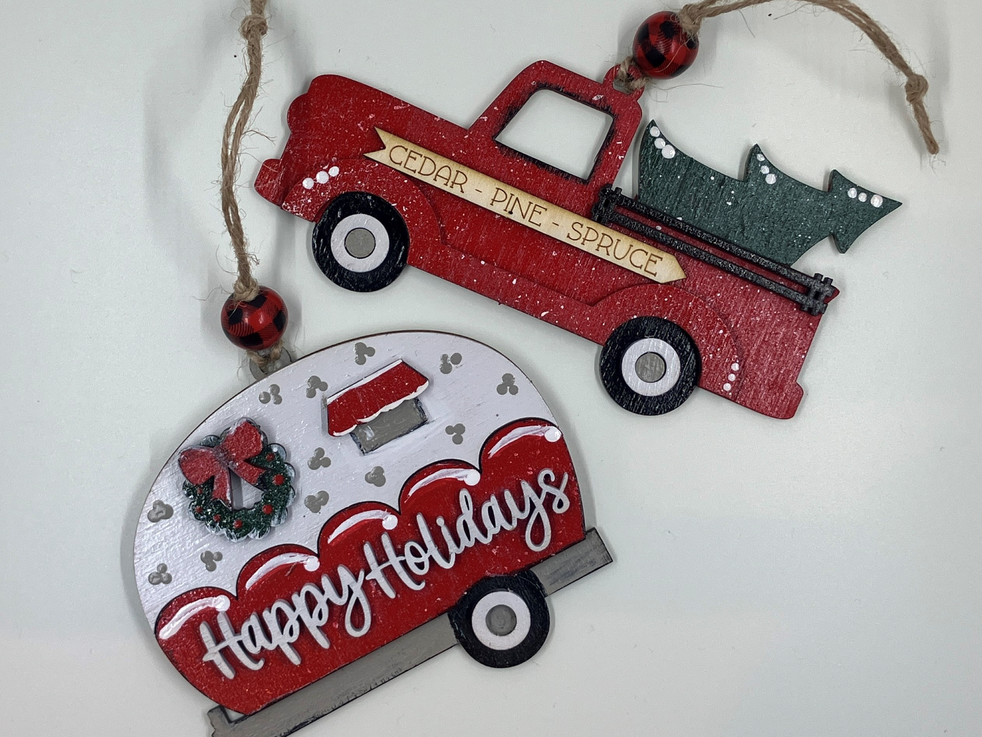 Two holiday ornaments are laid out on a table. One of them is a red pick up truck with a sign on it that reads "CEDAR - PINE - SPRUCE". A Christmas tree hangs out of the truck bed. The other ornament looks like a small red and white trailer with a wreath and "Happy Holidays" written across it.