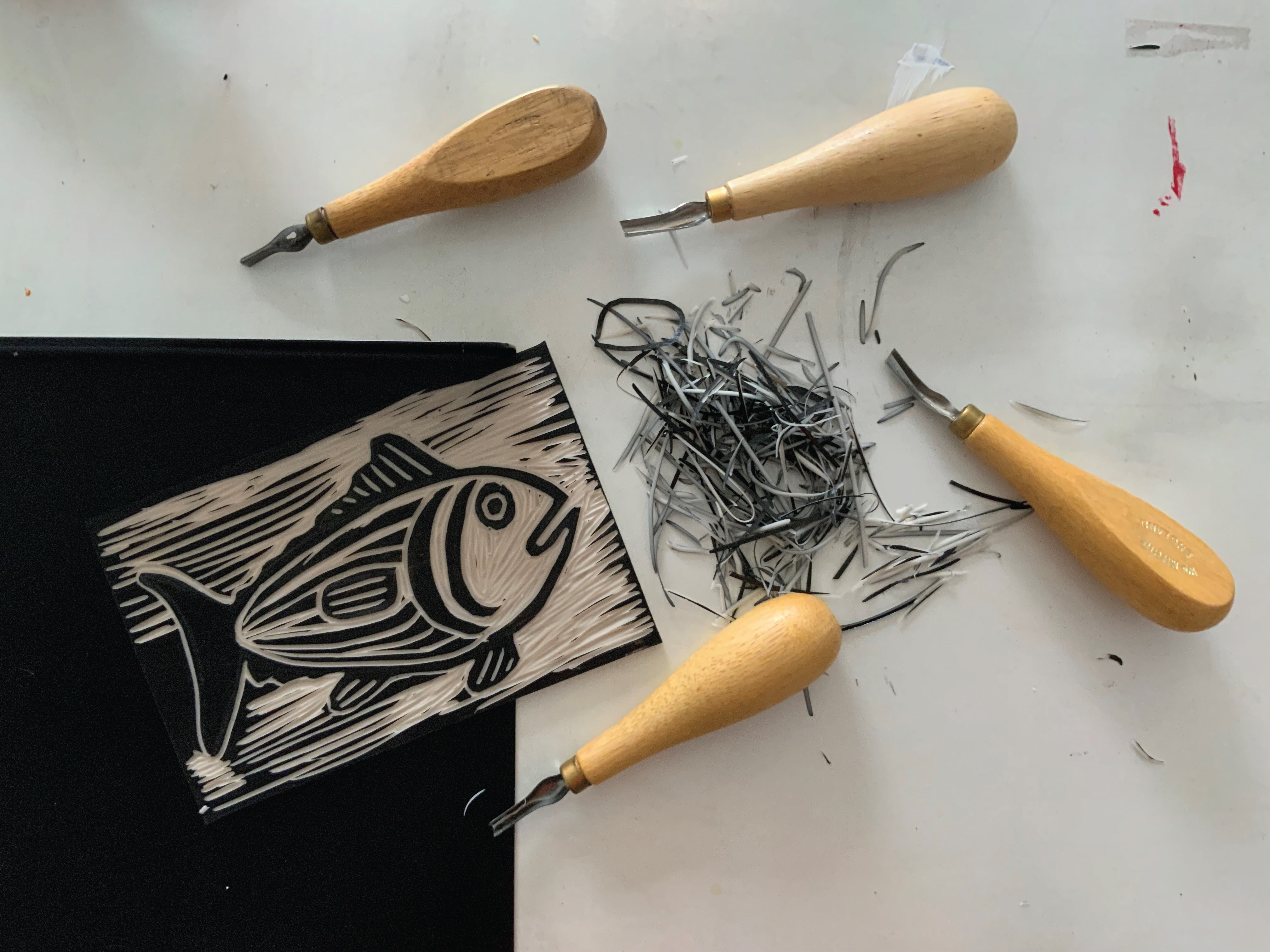 A linocut block featuring a stylized fish design is placed on a white surface surrounded by carving tools with wooden handles. Small shavings of linoleum are scattered nearby, showcasing the block printing process.