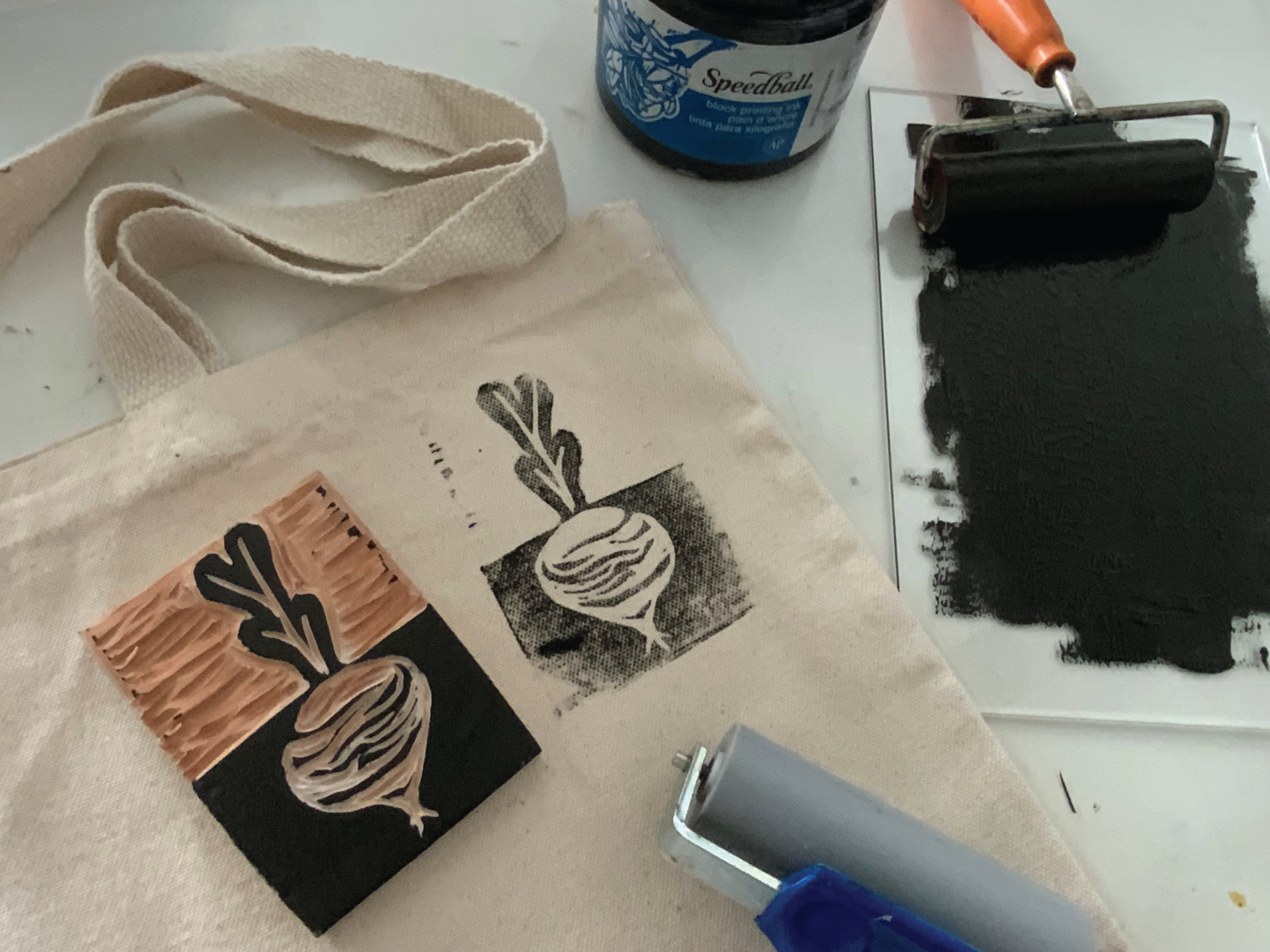 A linocut printing setup featuring a carved block with a beet design, black ink applied to a roller, and prints on a beige canvas tote bag. A jar of Speedball block printing ink and an inked plate are visible in the background.