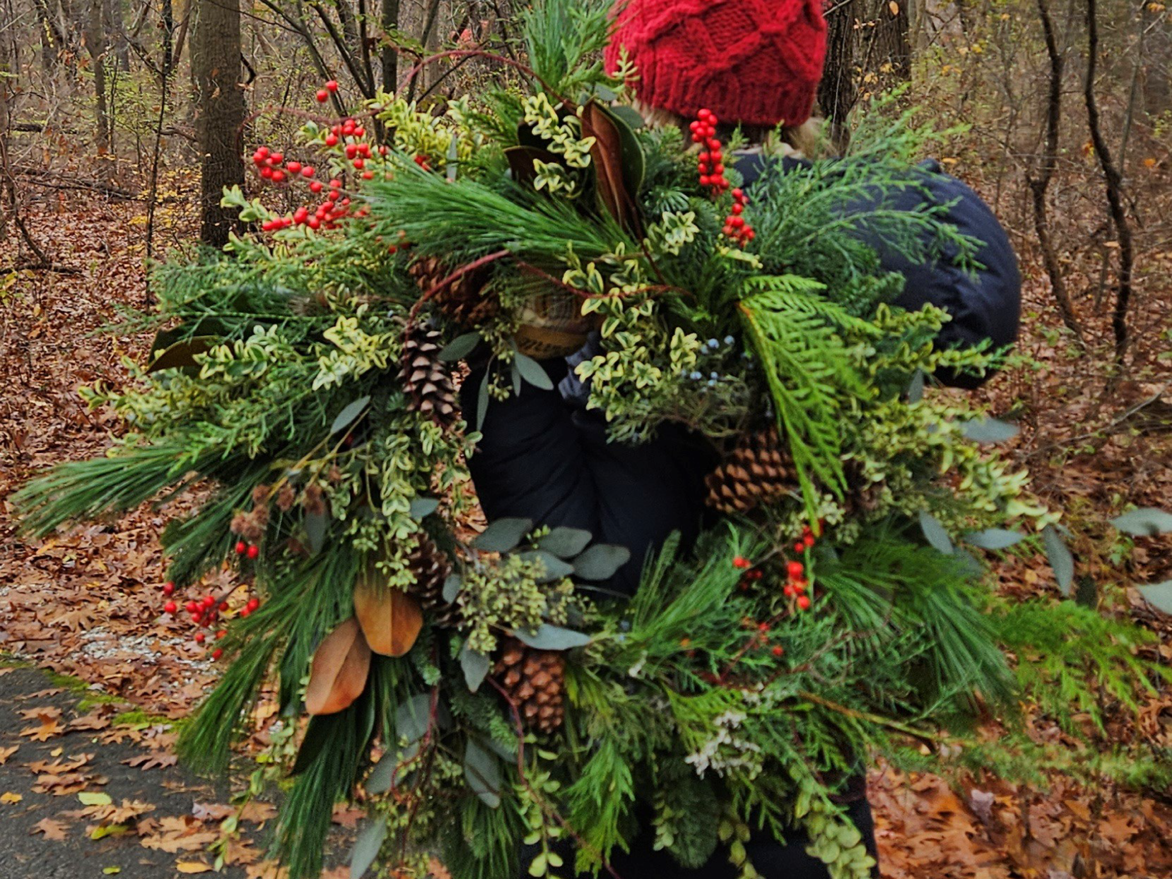 Someone wearing a bright red toque, holds up a wreath made of various winter greenery.