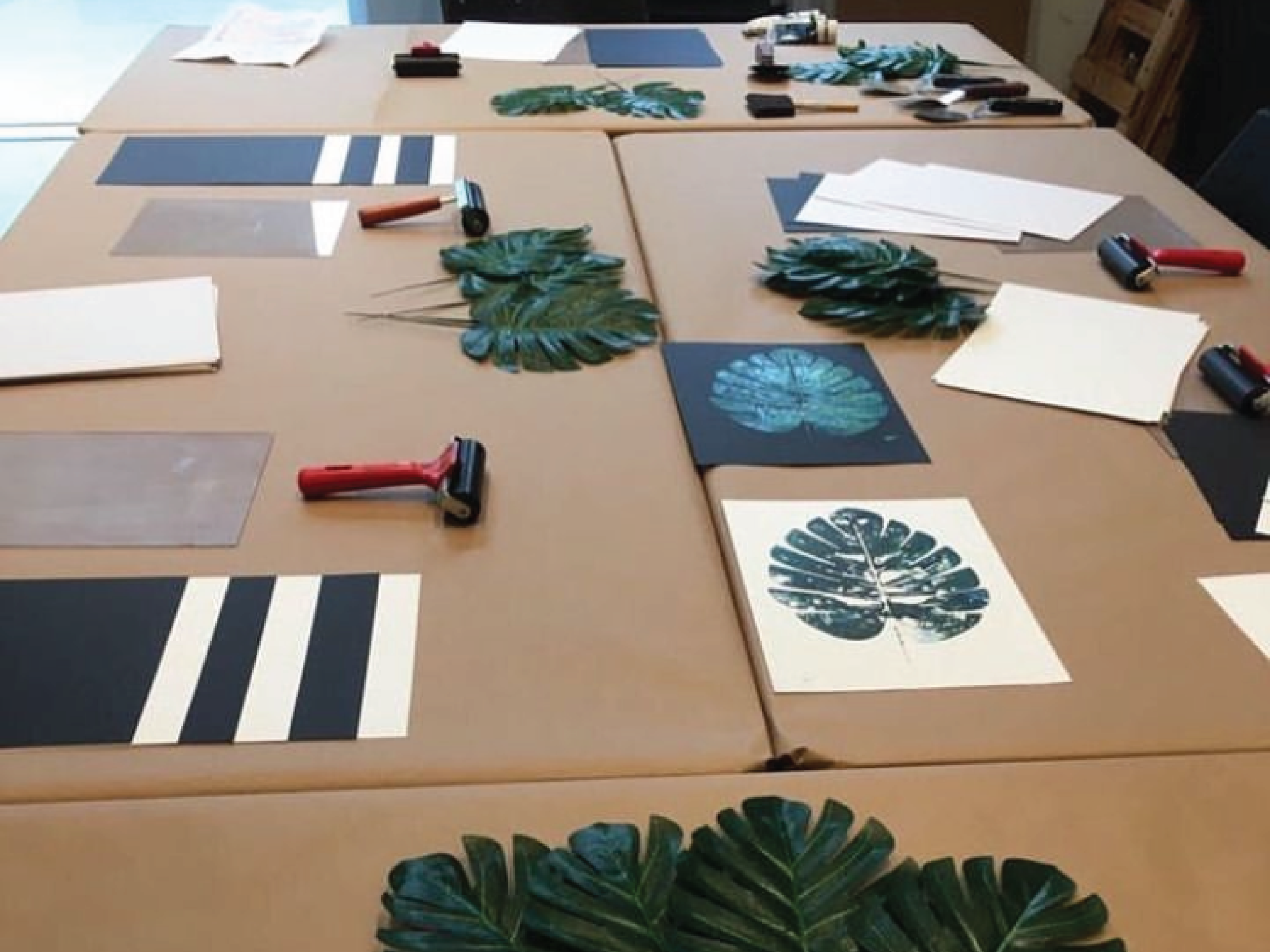 Workshop table covered in brown kraft paper, displaying supplies for botanical monoprinting. Large green leaves, ink rollers, black and white paper, and completed prints of leafy patterns are neatly arranged.