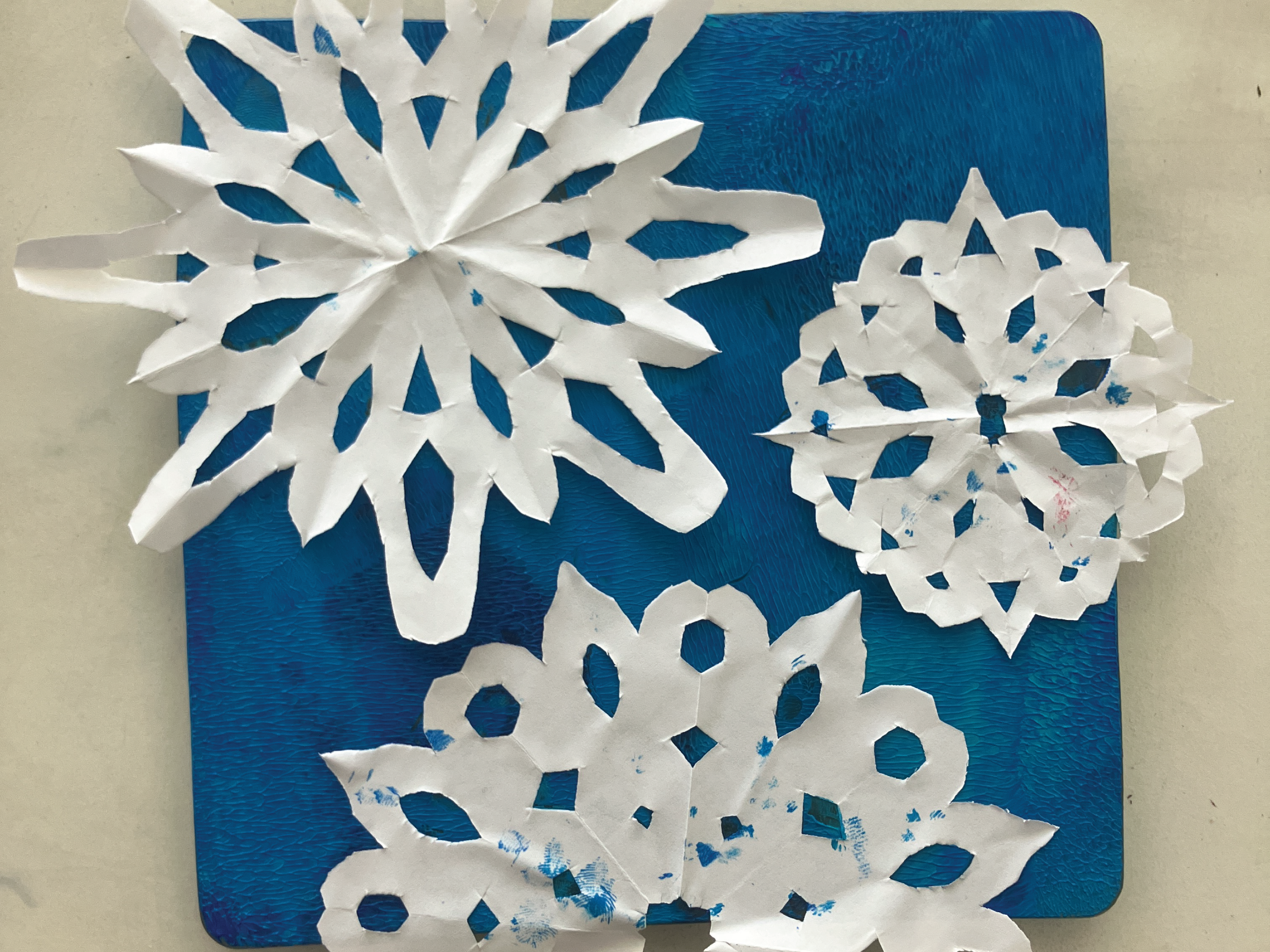 White paper snowflakes lay on top of a blue painted Gelli Plate.