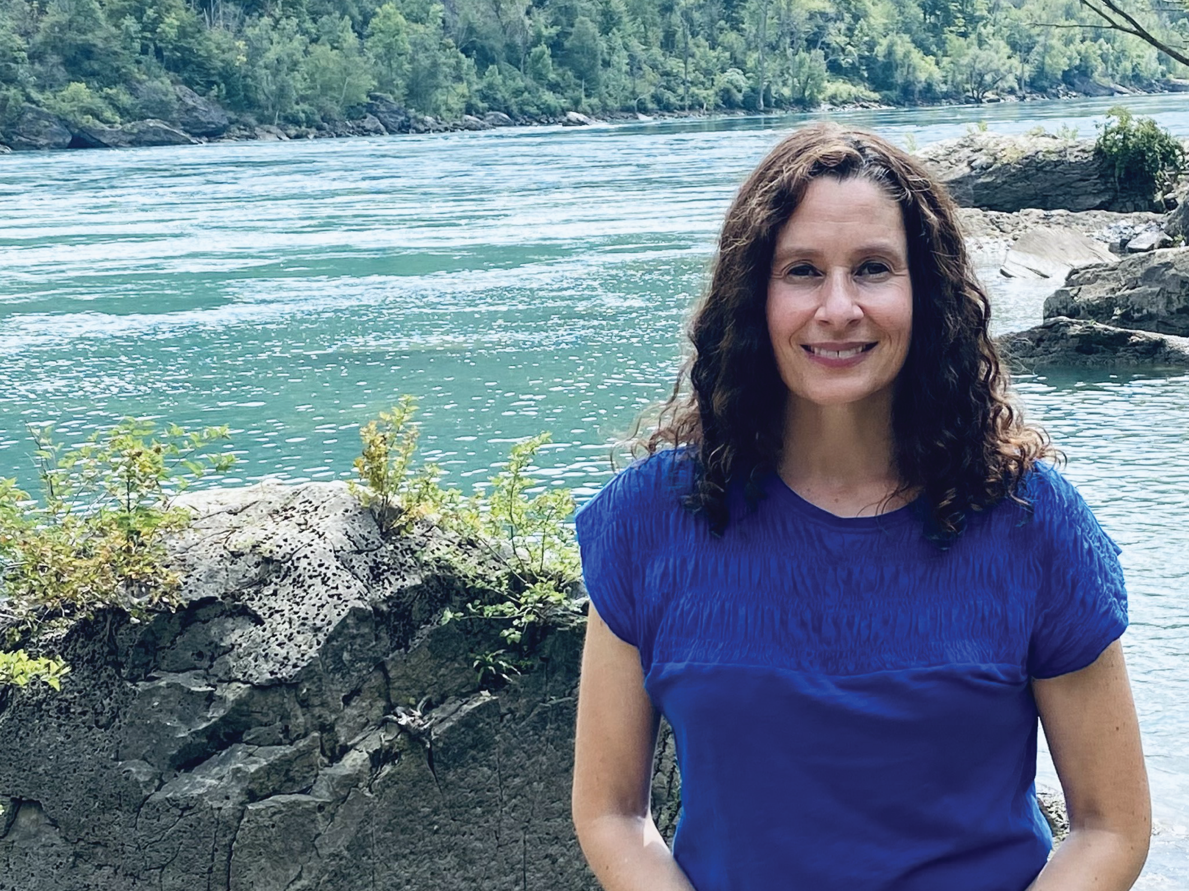 Professor Pauline Wakeham stands in front of a rocky lake wearing a deep blue t-shirt.