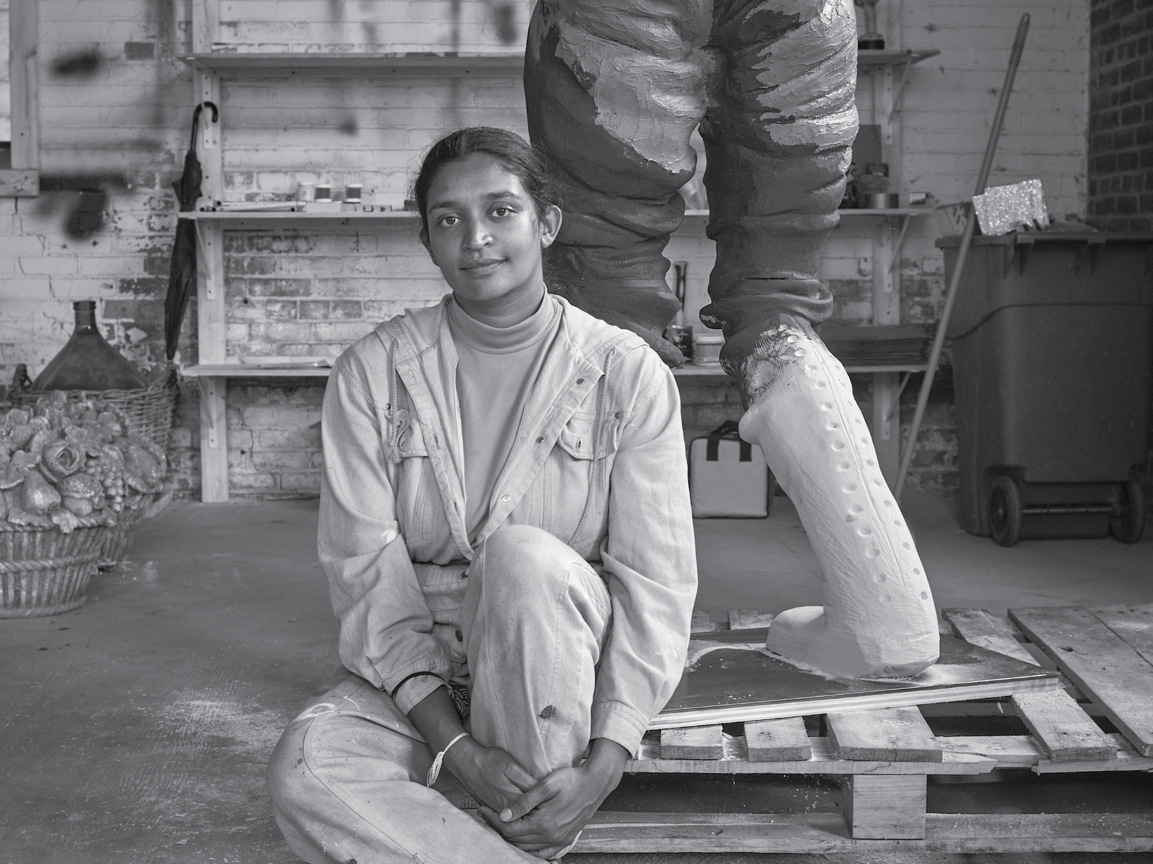 Artist Rajni Perera sits on a wooden pallet on the floor of an art studio. Shelves with assorted projects are visible in the background and a sculpture of what appears to be an arctic explorers legs accompany the artist on the wooden pallet. 