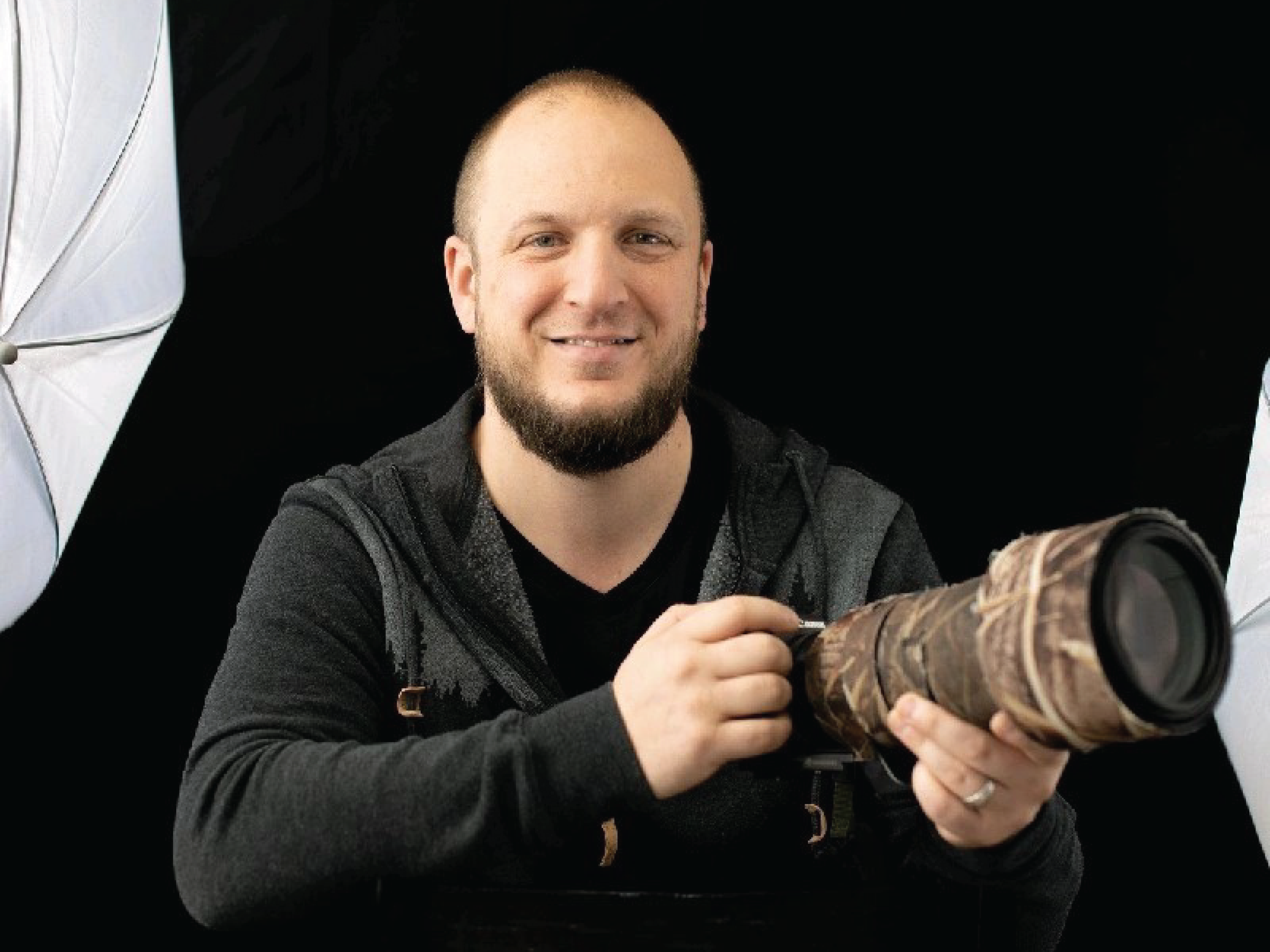 Photographer Shaun Antle poses with a camo outfitted, wide angle lens camera. He stands behind a black backdrop.