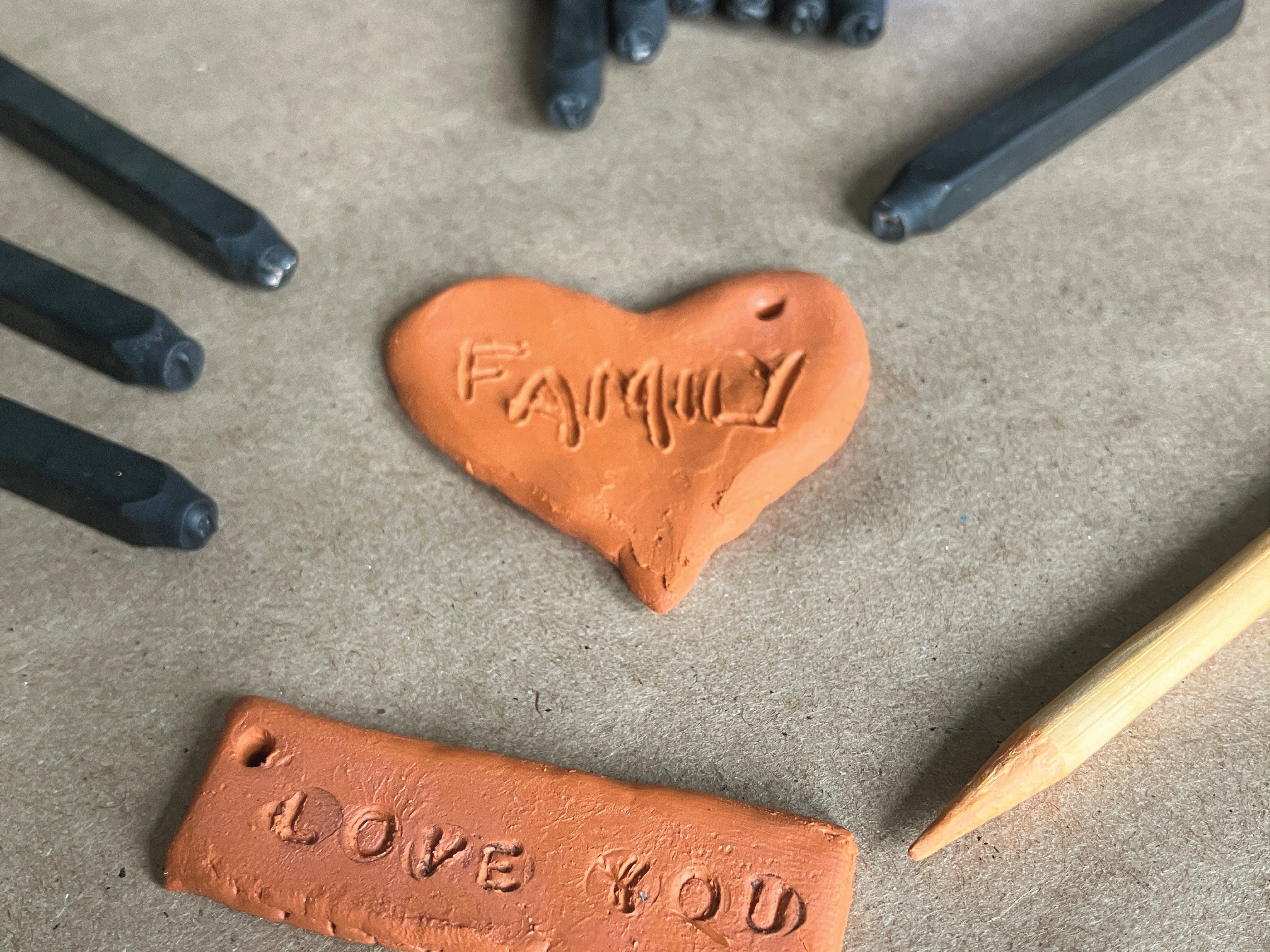 Two pieces of terracotta airdry clay, one in the shape of a heart and one in the shape of a rectangle, is surrounded by letter stamps and a pointed wooden pick. On the heart-shaped piece of clay, the word "Family" is etched into it. On the rectangle piece of clay, the words "love you" are stamped into it.