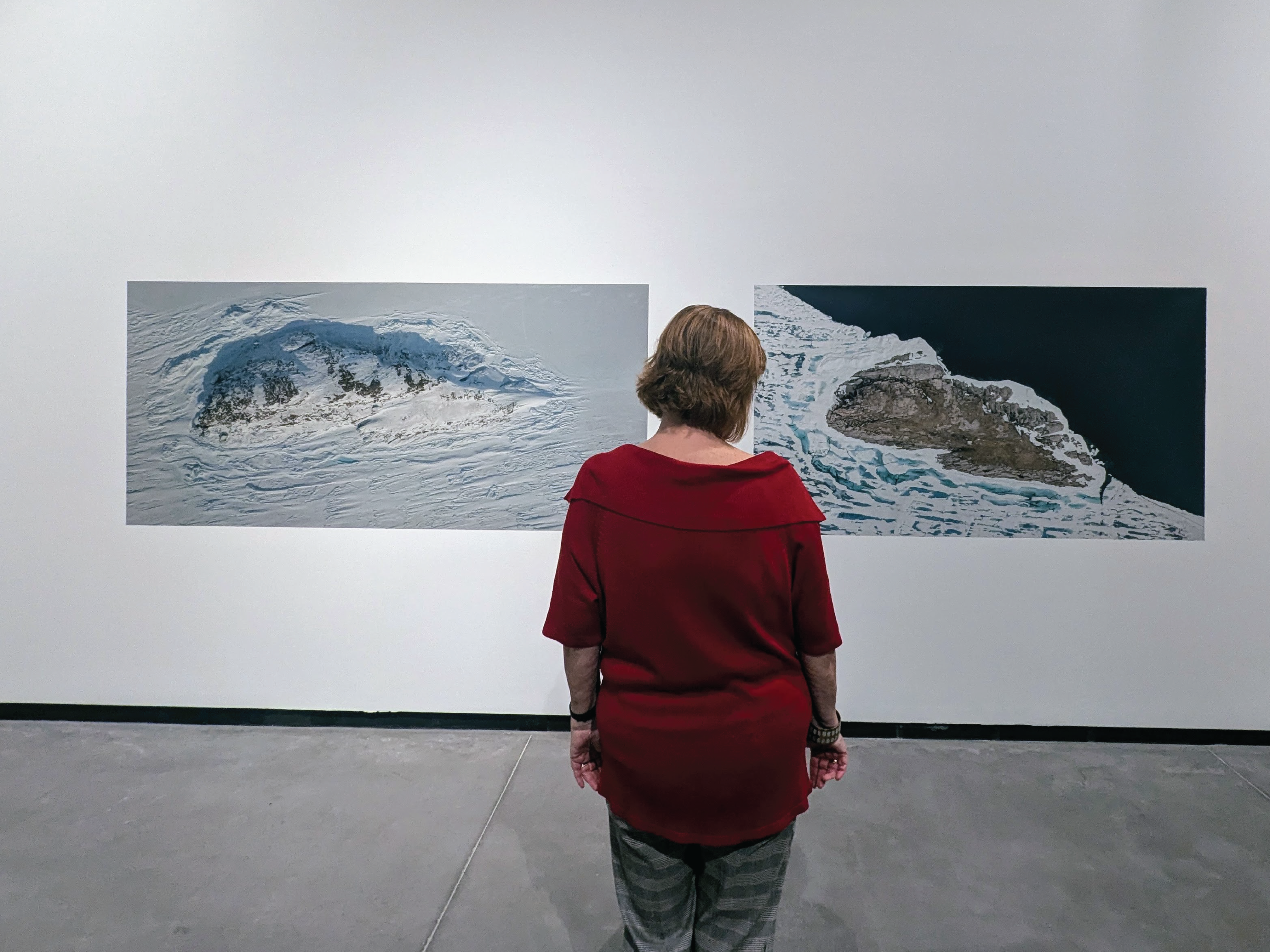 A women wearing a bright red shirt, stands in front of two large aerial photos of icebergs.