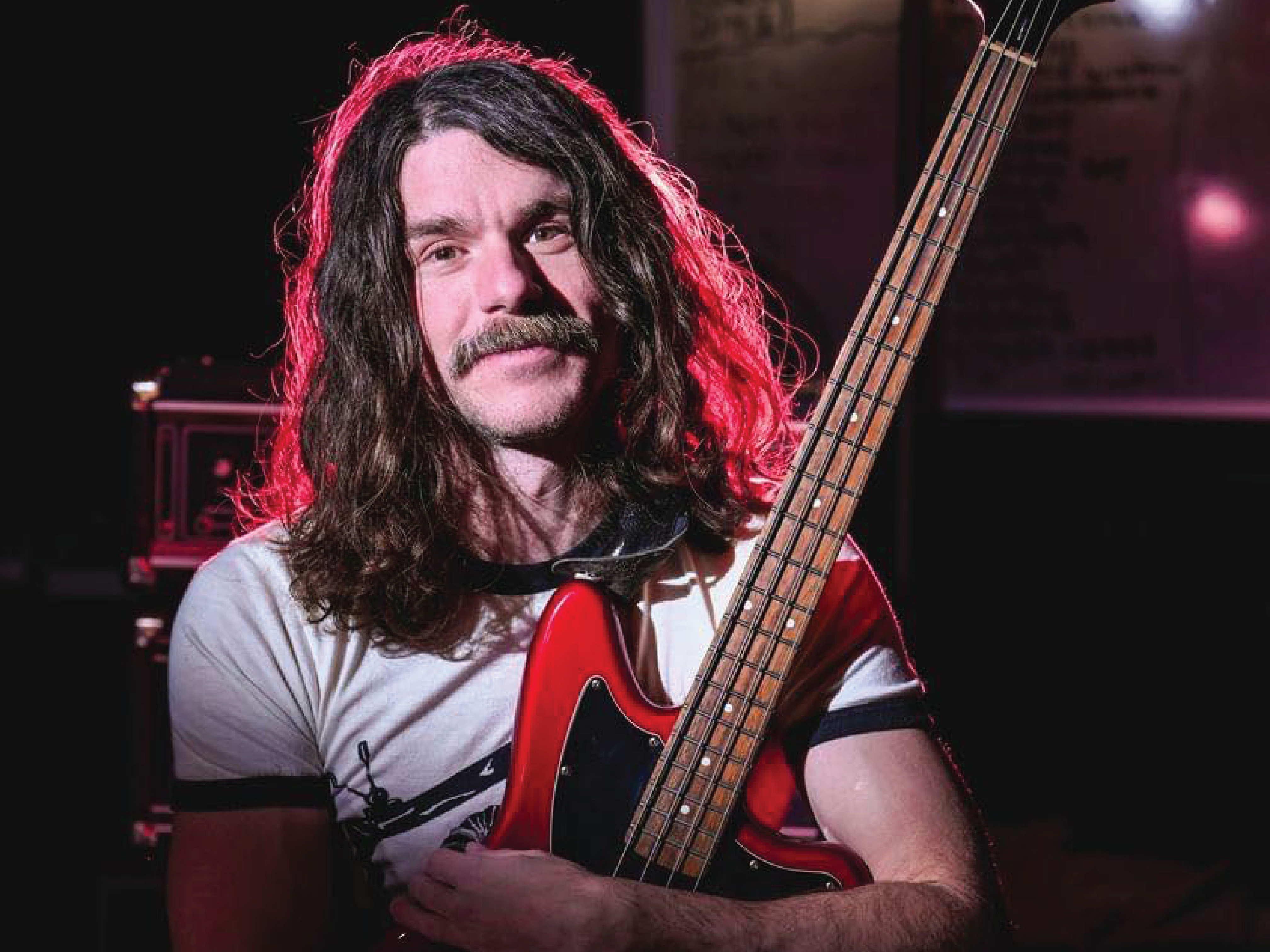 A gentleman with long, curly, brown hair is holding a black and red electric guitar and backlit with a red flood light, illuminating his hair to create a red halo like lighting effect.